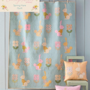 Può includere: Una trapunta blu chiaro con un motivo di coniglietti rosa e gialli e fiori. La trapunta è appesa a una parete blu chiaro. Ci sono due cuscini su una sedia davanti alla trapunta, uno è rosa e l'altro è giallo. I cuscini hanno lo stesso motivo della trapunta.