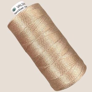 May include: A spool of light tan, metallic-toned thread wound in a cross-hatch pattern. A white label at the top reads VALDANI and Hand-Dyed, Cotton. The thread has a subtle sheen and is tightly coiled around the cylindrical core of the spool.