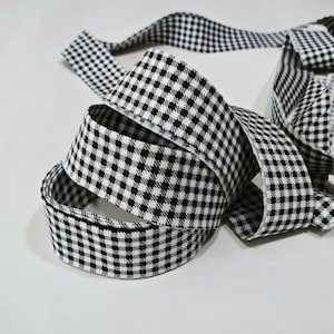 May include: A roll of fabric ribbon featuring a black and white gingham check pattern. The ribbon is coiled in a circular shape with a portion of the fabric extending outward, displaying the consistent small checkered design throughout the material.