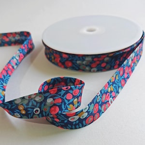 May include: A spool of fabric ribbon featuring a dark blue background with a repeating floral pattern in shades of bright pink, light blue, and muted yellow. The ribbon is wound around a white plastic core, with a portion unspooled and curled.