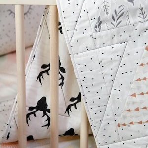 May include: A white and black baby quilt with a deer antler pattern and geometric designs. The quilt is draped over a crib, showcasing its soft texture and decorative appeal. The quilt features black polka dots and gray leaf patterns.