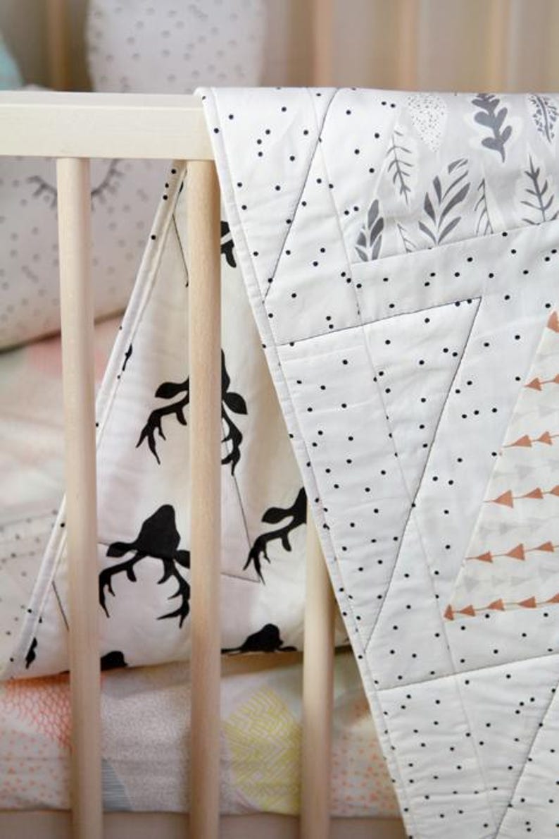 May include: A white and black baby quilt with a deer antler pattern and geometric designs. The quilt is draped over a crib, showcasing its soft texture and decorative appeal. The quilt features black polka dots and gray leaf patterns.