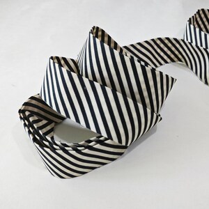 May include: A continuous length of fabric ribbon featuring a repeating pattern of diagonal black and beige stripes. The ribbon is folded into several overlapping loops, showcasing the consistent striped design across the entire length of the material.
