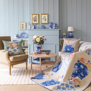 May include: A cozy living room scene with a light blue wall and wooden floor. The room features a beige sofa with a floral quilt and decorative pillows. A round pink table holds a vase of flowers. Artwork and blue and white ceramics adorn the room.