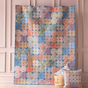 May include: A blue and pink floral quilt with a geometric pattern hangs on a pink wall. Two matching pillows are in front of the quilt.