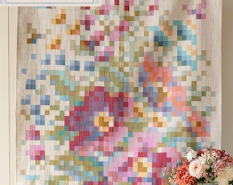 Op de afbeelding: Een grote, rechthoekige quilt met een bloemenontwerp. De quilt heeft een gepixeld patroon van bloemen in tinten roze, paars, blauw en geel tegen een neutrale achtergrond. De tekst "Tilda CHAMBRAY BASICS Flower Branch Embroidery Quilt" staat linksboven.