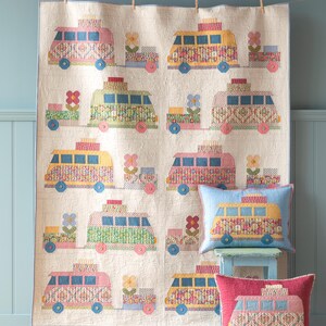 May include: A colorful quilt featuring a pattern of vintage camper vans with floral designs. The quilt is hanging on a light blue wall and there are two matching pillows in the foreground.