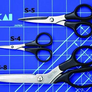 Może przedstawiać: Trzy pary nożyczek marki KAI z czarnymi uchwytami i srebrnymi ostrzami są pokazane na niebieskiej macie do cięcia. Nożyczki są oznaczone jako S-5, S-4 i S-8. Mata ma linie siatki i wymiary w cm.