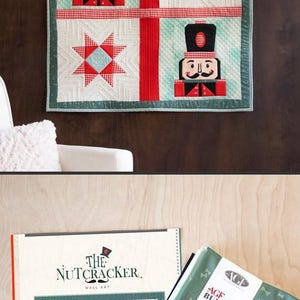 Puede incluir: Un tapiz acolchado cuadrado con dos figuras de cascanueces y dos patrones de estrellas en rojo, blanco y verde azulado. Debajo, un folleto de patrones titulado The Nutcracker Wall Art junto a una pila de telas plegadas en verde azulado.