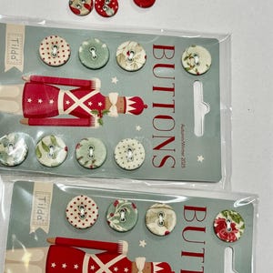 May include: A collection of decorative buttons in various designs. Some buttons feature red floral patterns, while others have green and white floral or dotted patterns. The packaging includes a red soldier graphic and the text "BUTTONS Autumn/Winter 2025".