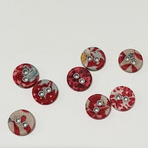 May include: A collection of round fabric buttons with a floral pattern in red, white, and gray. Each button has two holes for sewing and a metal rim. The buttons are scattered on a white surface.