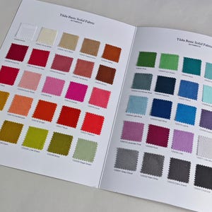 Peut inclure: Livre ouvert présentant des échantillons de tissus de différentes couleurs. Les pages affichent une gamme de couleurs unies, du jaune pâle au bleu marine, chaque échantillon étant étiqueté avec un code unique. Le texte "Tilda Basic Solid Fabric" est imprimé en haut de chaque page.