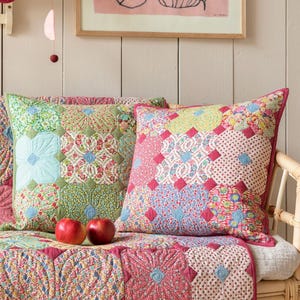 May include: Two decorative throw pillows with patchwork floral patterns in shades of pink, green, and blue. The pillows are on a bench with a matching quilt. Two red apples rest on the quilt.