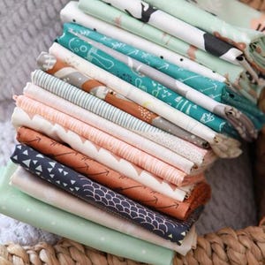 May include: A stack of folded fabric squares in various patterns and colors. Designs include woodland animals, arrows, and geometric shapes. Colors range from teal and coral to navy and mint green. The fabrics are neatly arranged in a woven basket.