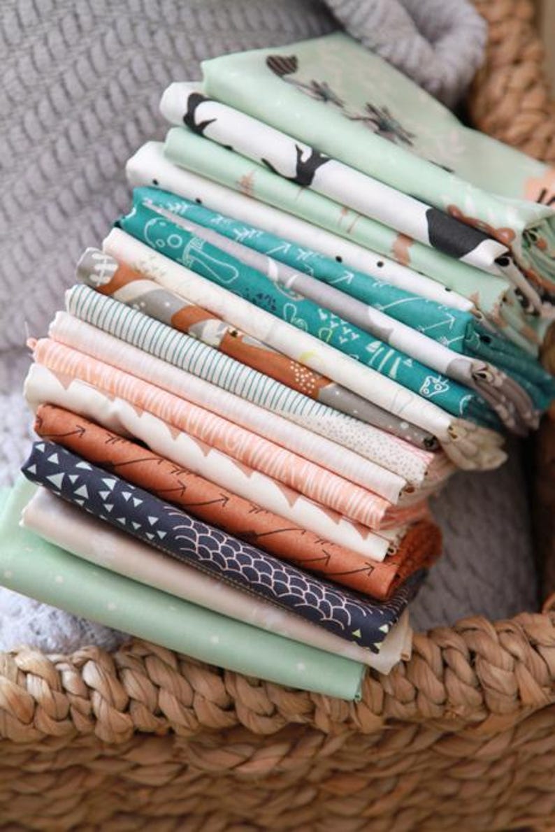 May include: A stack of folded fabric squares in various patterns and colors. Designs include woodland animals, arrows, and geometric shapes. Colors range from teal and coral to navy and mint green. The fabrics are neatly arranged in a woven basket.