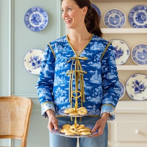 May include: A quilted jacket in blue and white featuring a toile-style pattern of elephants and pagodas, finished with olive green trim and front tie closures. The jacket is worn with blue denim jeans and paired with a two-tiered serving stand.