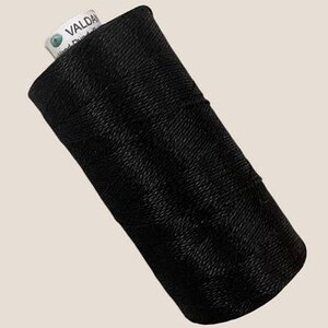 May include: A spool of black thread wound in a cross-hatch pattern. The top of the spool features a white label with the text VALDANI and Hand Dyed printed in black ink. The thread has a slight sheen and is tightly coiled around the cylindrical core.