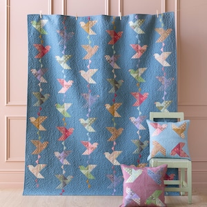 May include: A blue quilt with a pattern of colorful birds hanging on a pink wall. Two patterned throw pillows are on a light green stool.