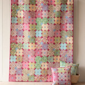 May include: A pink, green, and blue floral quilt with a quilted pattern of flowers. The quilt is hanging on a white wall and there are three matching floral pillows in front of it.