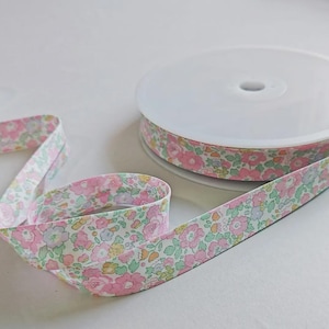 May include: A spool of fabric ribbon featuring a repeating floral pattern in shades of pink, light green, and soft yellow. The ribbon is wound around a white plastic circular reel, with a portion of the patterned fabric extending outward.