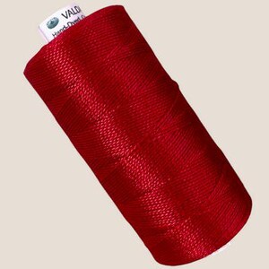 Pode incluir: Um carretel de linha vermelha vibrante enrolado firmemente em um padrão cruzado. O topo do carretel apresenta uma etiqueta branca com o texto VALDANI e Hand-Dyed, exibindo um pequeno logotipo circular. A linha tem uma textura lisa e brilhante.