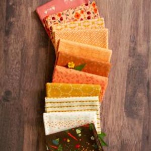 May include: A stack of folded fabric squares in a gradient of colours ranging from coral and orange to yellow, cream, and dark brown. The fabrics feature various patterns including small flowers, geometric shapes, dots, and a vegetable print.