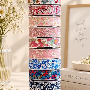 May include: A vertical stack of twelve spools of fabric ribbon, each featuring a distinct floral pattern in various colors including pink, blue, red, and yellow. The ribbons are arranged in a column on a circular base, showcasing diverse botanical prints.