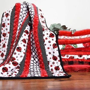May include: A red, black, and white quilt with a ladybug pattern. The quilt is folded and displayed with a stack of red, white, and black fabric squares.