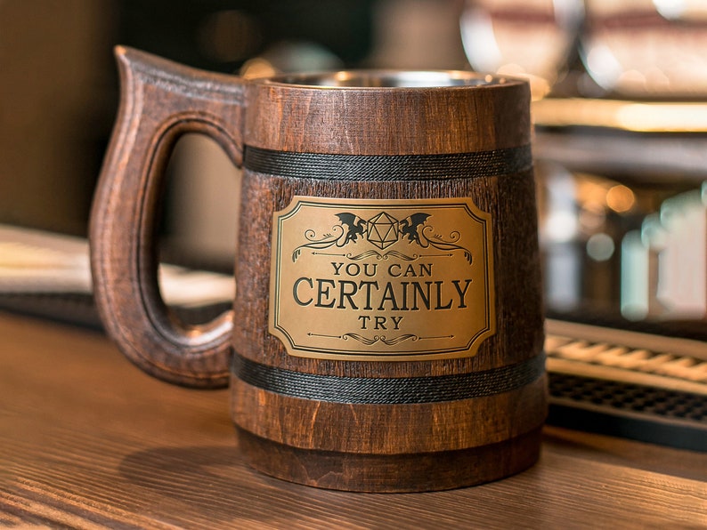 May include: A wooden beer stein with a black band and a gold metal plaque that reads "You Can Certainly Try". The plaque features a black and white illustration of a dragon and a 20-sided die.