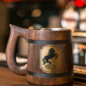May include: A wooden beer mug with a large handle and a metal interior. The mug features a brass emblem with a black horse and the words "Prancing Pony". The wood has a dark brown finish.