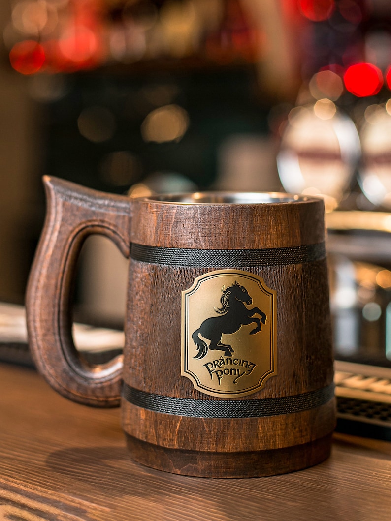 May include: A wooden beer mug with a large handle and a metal interior. The mug features a brass emblem with a black horse and the words "Prancing Pony". The wood has a dark brown finish.