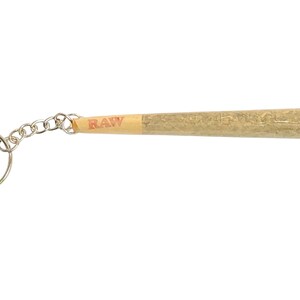 May include: A silver keychain with a realistic-looking, brown, rolled-up joint with a RAW label. The joint is attached to a silver chain and a silver ring.