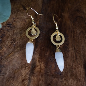 Könnte beinhalten: Ein Paar goldene Ohrringe mit einem weißen tropfenförmigen Stein und einem goldenen Kreisanhänger mit einem Muschelmotiv.