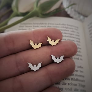 Könnte beinhalten: Ein Paar Ohrringe mit einem Fledermaus-Design. Ein Paar ist golden und das andere ist silber.