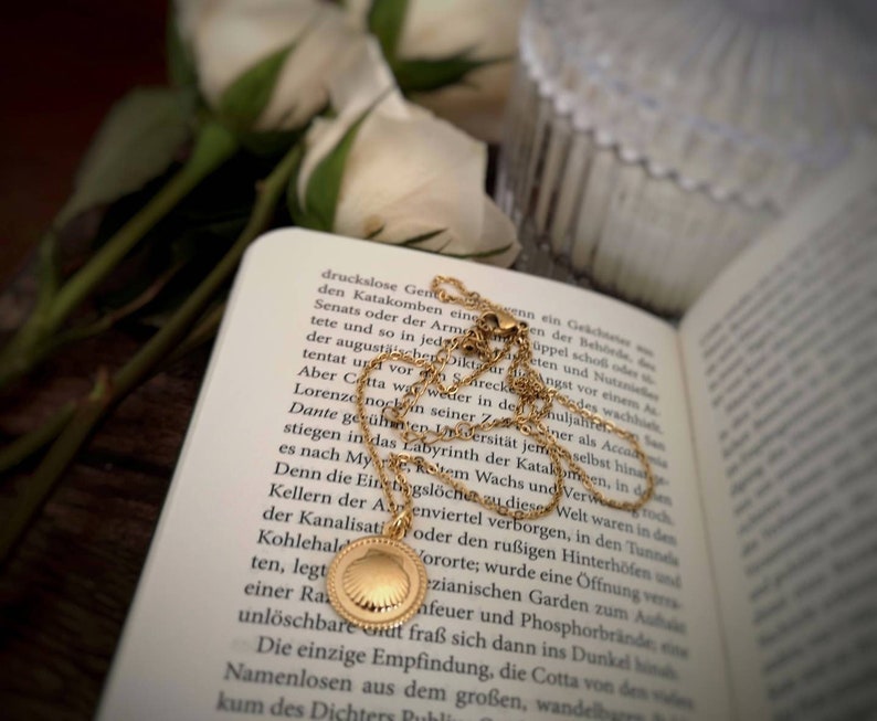 May include: A gold chain necklace with a seashell pendant. The necklace is lying on a book with white flowers in the background.