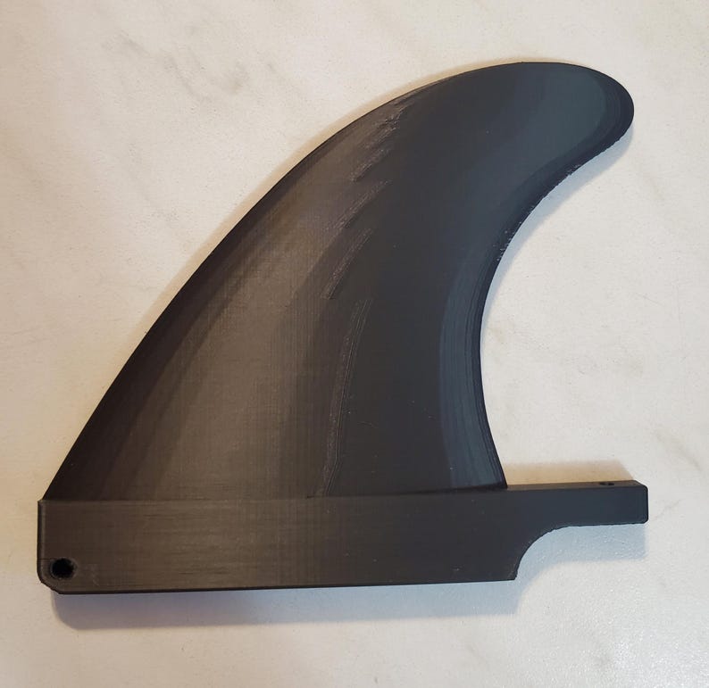 Pode incluir: Uma barbatana de prancha de surf preta, com uma l&acirc;mina curva e uma base plana com um orif&iacute;cio para fixa&ccedil;&atilde;o. A barbatana &eacute; feita de um material preto fosco e &eacute; projetada para esportes aqu&aacute;ticos.