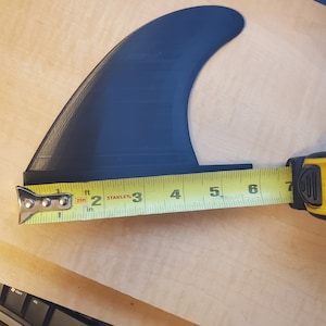 Pode incluir: Uma barbatana de prancha de surf preta &eacute; medida com uma fita m&eacute;trica amarela. A barbatana tem aproximadamente 15 cm de comprimento. A barbatana &eacute; feita de um material escuro e s&oacute;lido e &eacute; projetada para uma prancha de surf.