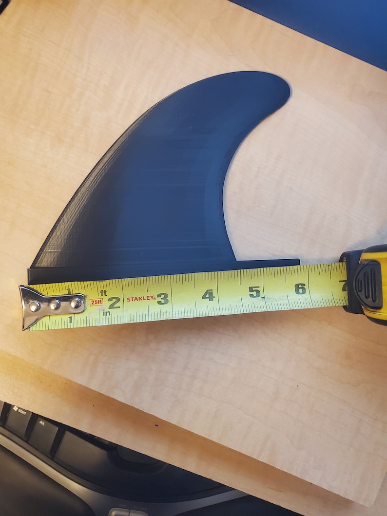 Pode incluir: Uma barbatana de prancha de surf preta &eacute; medida com uma fita m&eacute;trica amarela. A barbatana tem aproximadamente 15 cm de comprimento. A barbatana &eacute; feita de um material escuro e s&oacute;lido e &eacute; projetada para uma prancha de surf.