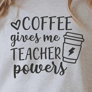 May include: A light gray sweatshirt with the text "COFFEE gives me TEACHER powers" in black. The design includes a coffee cup with a lightning bolt. The person is wearing light blue jeans.