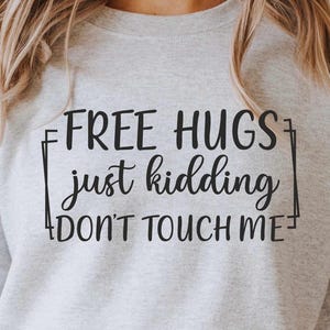 以下が含まれることがあります： 「FREE HUGS just kidding DON'T TOUCH ME」の黒い文字がプリントされたライトグレーのスウェットシャツ。 ライトブルーのデニムジーンズとボタンフライが組み合わされています。