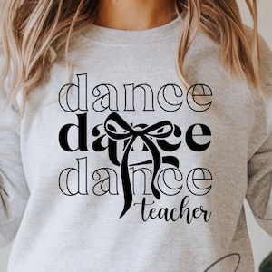 May include: A light gray sweatshirt with the words "dance dance dance teacher" in a black, outlined font. A black bow graphic is between the words "dance". The model is wearing blue jeans.