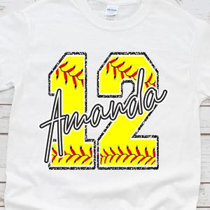 May include: White t-shirt with a softball-themed design. The design features the number 12 in yellow with black outlines and red stitching, the name "Amanda" in a script font, and a softball texture.