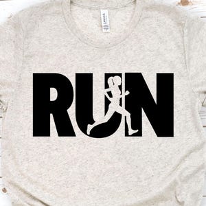 May include: A light beige t-shirt with the word "RUN" in large black letters. The letter "U" is replaced by a silhouette of a person running. The shirt is made of a soft material and has short sleeves.