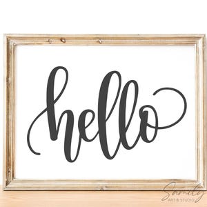 May include: A framed print with the word "hello" in elegant black script. The artwork is set against a white background and framed in a light brown wooden frame. The overall aesthetic is simple and modern.