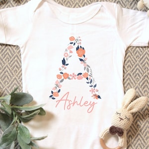 May include: White baby onesie with a floral letter "A" design in coral, navy, and blush tones. The name "Ashley" is written below the letter in a matching blush color. A crochet bunny rattle is next to the onesie.
