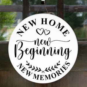 Puede incluir: Un letrero redondo de madera blanca con letras negras. El letrero dice "NEW HOME, NEW BEGINNING, NEW MEMORIES" con dos corazones y un diseño de hojas decorativas. Un regalo de inauguración.