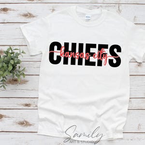 May include: White t-shirt with the word "CHIEFS" in large black block letters. The words "kansas city" are in red script font, centered below the word "CHIEFS". The shirt is displayed on a light wood surface.