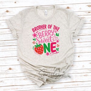 May include: A heather gray short-sleeve t-shirt featuring a graphic print with the text BROTHER OF THE BERRY SWEET ONE in pink, red, and green. The design includes a red strawberry, small pink flowers, green leaves, and red heart outlines.