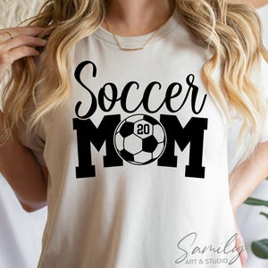 May include: Cream-colored t-shirt with the words "Soccer Mom" in black script and block letters. A soccer ball graphic is in the center of the word "MOM", with the number "20" inside the ball. The shirt is from Sami Art & Studio.
