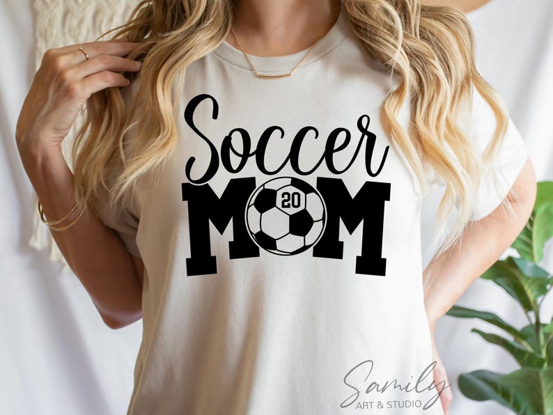 May include: Cream-colored t-shirt with the words "Soccer Mom" in black script and block letters. A soccer ball graphic is in the center of the word "MOM", with the number "20" inside the ball. The shirt is from Sami Art & Studio.
